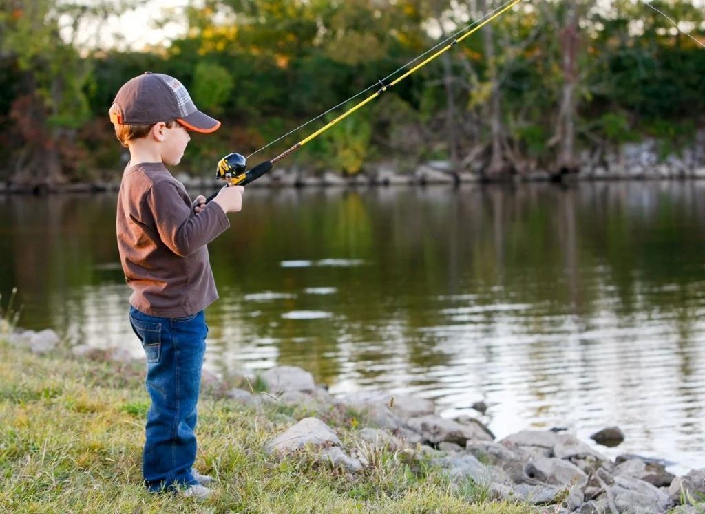 Child holding a lightweight kids fishing rod while fishing by a calm lake with adult supervision